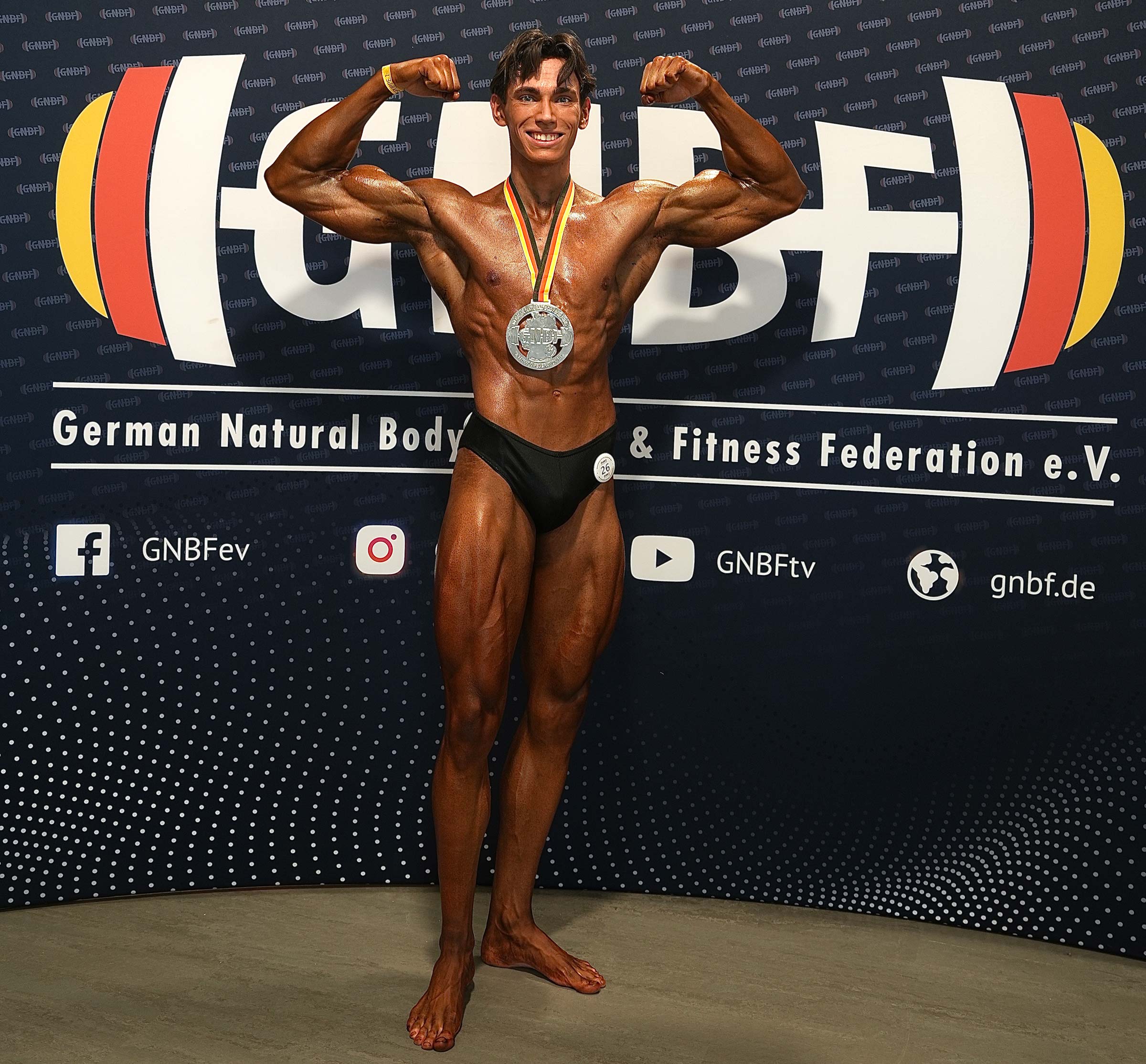 Trainer posiert auf der Bühne, zeigt seinen muskulösen Körper und trägt eine Medaille um den Hals.